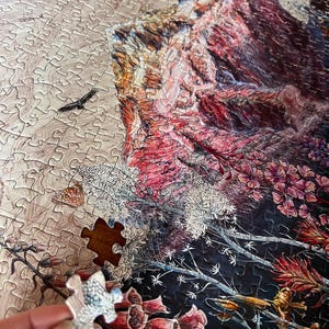 Pu&ograve; includere: Un puzzle parzialmente completato con una scena paesaggistica dettagliata. Il puzzle presenta una montagna, fiori e un uccello. Un singolo pezzo del puzzle viene tenuto in mano, pronto per essere posizionato. La tavolozza dei colori include sfumature di rosso, rosa, marrone e bianco.