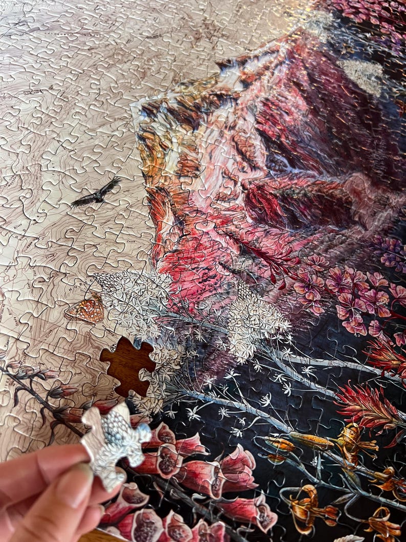 Pu&ograve; includere: Un puzzle parzialmente completato con una scena paesaggistica dettagliata. Il puzzle presenta una montagna, fiori e un uccello. Un singolo pezzo del puzzle viene tenuto in mano, pronto per essere posizionato. La tavolozza dei colori include sfumature di rosso, rosa, marrone e bianco.
