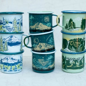 May include: A stack of six enamel mugs with different designs featuring national parks. The mugs are blue, green, and white with illustrations of mountains, trees, and water. The text on the mugs includes "Mount Rainier National Park", "Olympic National Park", and "Mount Rainier National Park".