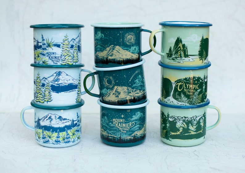 May include: A stack of six enamel mugs with different designs featuring national parks. The mugs are blue, green, and white with illustrations of mountains, trees, and water. The text on the mugs includes "Mount Rainier National Park", "Olympic National Park", and "Mount Rainier National Park".