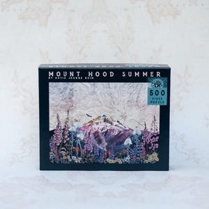Pu&ograve; includere: Una scatola nera con un puzzle da 500 pezzi. L'immagine del puzzle raffigura il Monte Hood in un paesaggio estivo, con fiori colorati in primo piano. Il testo "MOUNT HOOD SUMMER" &egrave; stampato in alto.