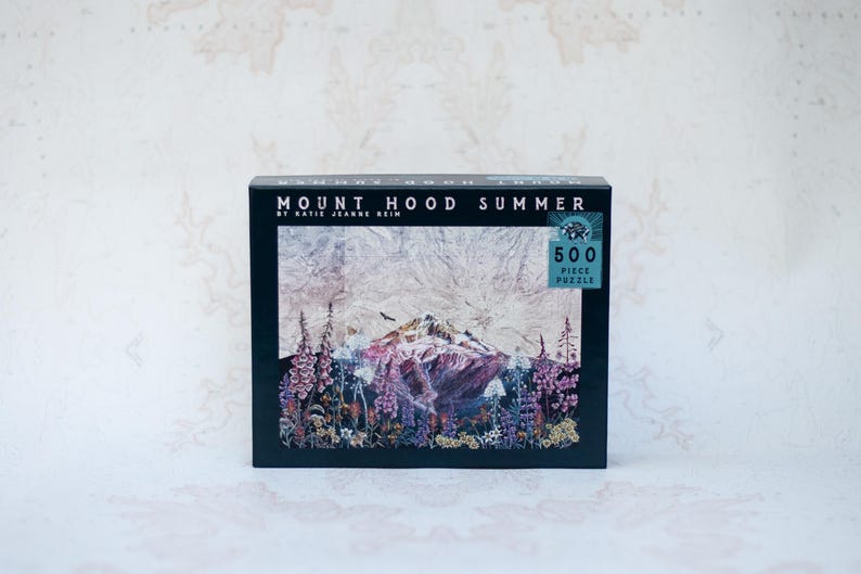Pu&ograve; includere: Una scatola nera con un puzzle da 500 pezzi. L'immagine del puzzle raffigura il Monte Hood in un paesaggio estivo, con fiori colorati in primo piano. Il testo "MOUNT HOOD SUMMER" &egrave; stampato in alto.