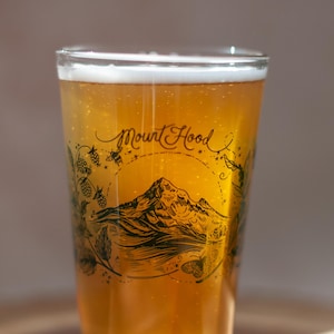 May include: A clear glass pint glass filled with a golden amber liquid and white foam. The glass features a black ink illustration of Mount Hood, surrounded by botanical elements like hops and leaves, with the text Mount Hood written above.