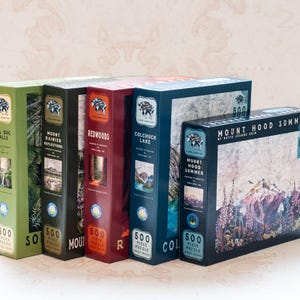 May include: A collection of five 500-piece jigsaw puzzles. The boxes are various colors, including green, black, red, and blue, each featuring landscape scenes. Titles include "Sol Duc Falls," "Mount Rainier Reflections," and "Mount Hood Summer."