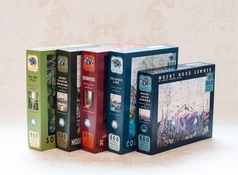 May include: A collection of five 500-piece jigsaw puzzles. The boxes are various colors, including green, black, red, and blue, each featuring landscape scenes. Titles include "Sol Duc Falls," "Mount Rainier Reflections," and "Mount Hood Summer."