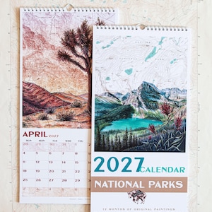 May include: A 2027 National Parks calendar by Katie Jeanne Reim featuring original paintings of landscapes. The calendar displays a Joshua Tree scene for April and a mountain lake scene on the cover, with spiral binding and printed dates.