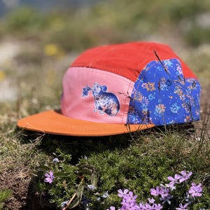 May include: A five-panel cap featuring a red crown, a flat orange brim, and a pink side panel with a blue graphic of a small rodent holding a flower. The back panel is blue with a repeating floral pattern in orange and light blue colors.