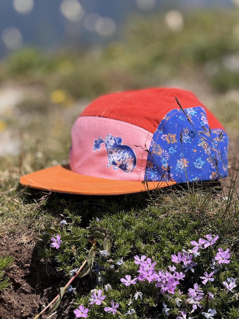 May include: A five-panel cap featuring a red crown, a flat orange brim, and a pink side panel with a blue graphic of a small rodent holding a flower. The back panel is blue with a repeating floral pattern in orange and light blue colors.