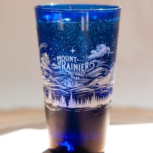 May include: A deep blue glass tumbler featuring a white etched design of Mount Rainier National Park. The graphic includes a star, swirling clouds, and a forest silhouette. The glass has a tapered shape and a smooth, translucent finish.
