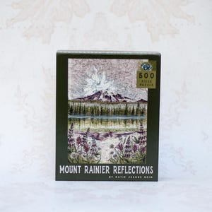 May include: A boxed 500-piece puzzle featuring a landscape scene of Mount Rainier reflected in a lake. The image shows a mountain with a snowy peak, trees, and purple flowers. The box is dark green with the text "Mount Rainier Reflections".
