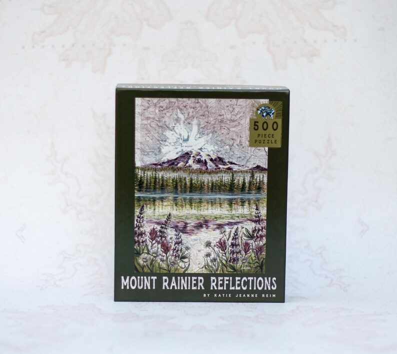 May include: A boxed 500-piece puzzle featuring a landscape scene of Mount Rainier reflected in a lake. The image shows a mountain with a snowy peak, trees, and purple flowers. The box is dark green with the text "Mount Rainier Reflections".