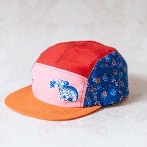 May include: A five-panel cap featuring a red top panel, a light pink front panel with a blue graphic of a small rodent holding a plant, a flat orange brim, and blue side panels decorated with a small, repeating floral pattern in orange and white.