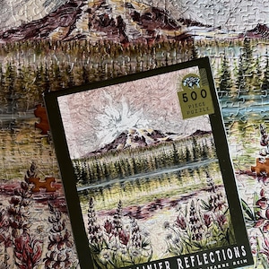 May include: A 500-piece puzzle featuring a landscape of Mount Rainier with a lake and trees. The puzzle box, with the text "Mount Rainier Reflections," sits on top of the partially assembled puzzle. The color palette includes greens, browns, and purples.