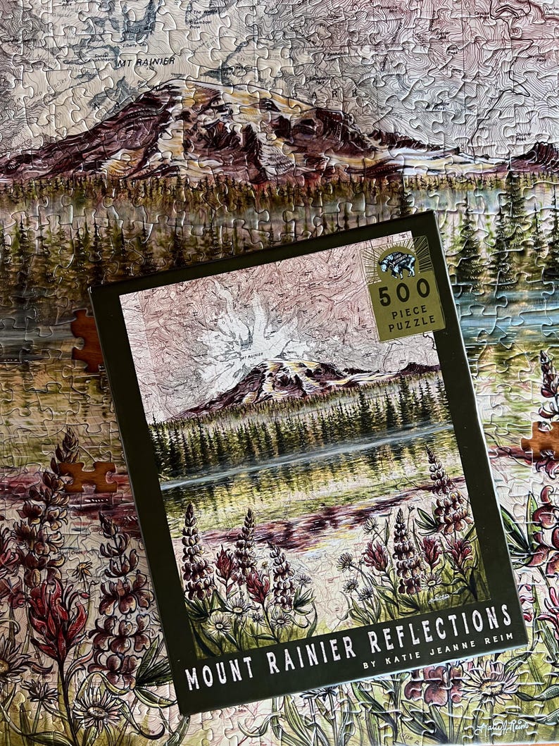 May include: A 500-piece puzzle featuring a landscape of Mount Rainier with a lake and trees. The puzzle box, with the text "Mount Rainier Reflections," sits on top of the partially assembled puzzle. The color palette includes greens, browns, and purples.