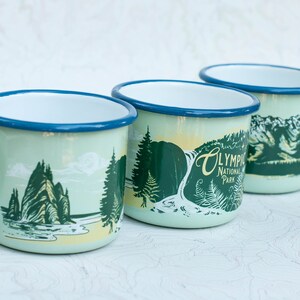 May include: Three enamel mugs with a light blue rim and a light green body. Each mug features a different illustration of a landscape from Olympic National Park. The text "Olympic National Park" is printed on one of the mugs.