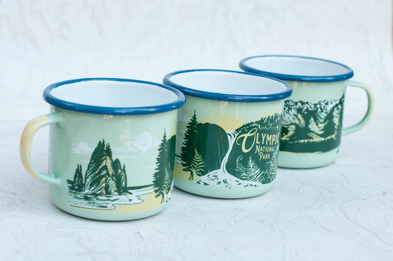 May include: Three enamel mugs with a light blue rim and a light green body. Each mug features a different illustration of a landscape from Olympic National Park. The text "Olympic National Park" is printed on one of the mugs.