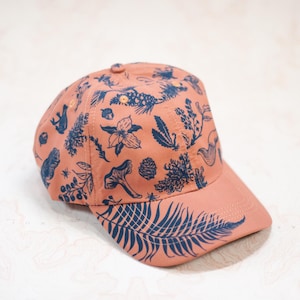 May include: A coral-colored baseball cap featuring a navy blue botanical pattern. The design includes illustrations of flowers, ferns, and mushrooms. The cap has a curved brim and a button on top.