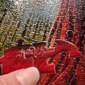 May include: A partially assembled jigsaw puzzle with a detailed image of a redwood tree. A red puzzle piece with the text "Coyote" and a signature is being placed. The puzzle pieces are silver.