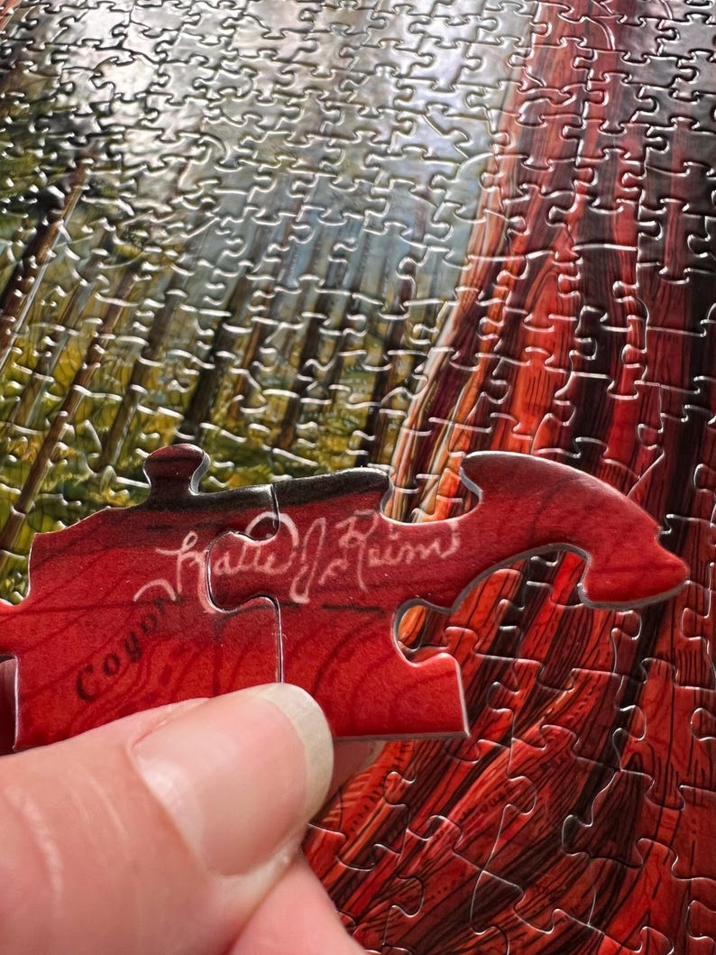 May include: A partially assembled jigsaw puzzle with a detailed image of a redwood tree. A red puzzle piece with the text "Coyote" and a signature is being placed. The puzzle pieces are silver.