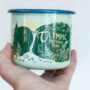 May include: A green and white enamel mug with a blue rim. The mug features a graphic of a waterfall in a forest with the text "Olympic National Park" in yellow.