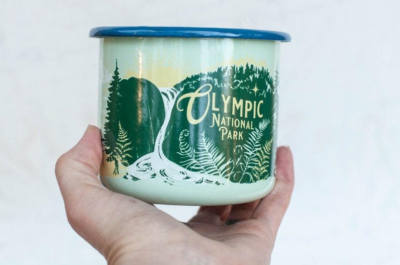 May include: A green and white enamel mug with a blue rim. The mug features a graphic of a waterfall in a forest with the text "Olympic National Park" in yellow.