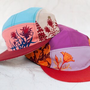 May include: Two colorful five-panel caps with floral designs. One cap has a light blue top, pink front panel with red flowers, and a burgundy brim. The other cap has a purple top, orange front panel with an orange flower, and a red brim.