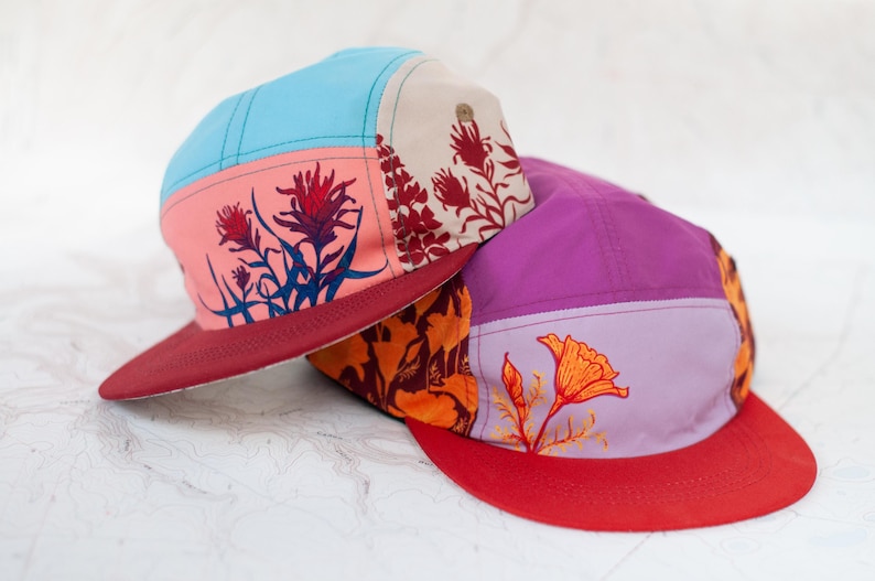 May include: Two colorful five-panel caps with floral designs. One cap has a light blue top, pink front panel with red flowers, and a burgundy brim. The other cap has a purple top, orange front panel with an orange flower, and a red brim.