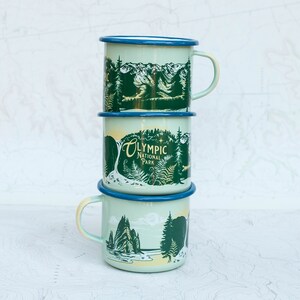 May include: A stack of three enamel camping mugs with a green and blue design. The top mug features a mountain scene, the middle mug says "Olympic National Park", and the bottom mug features a forest scene.