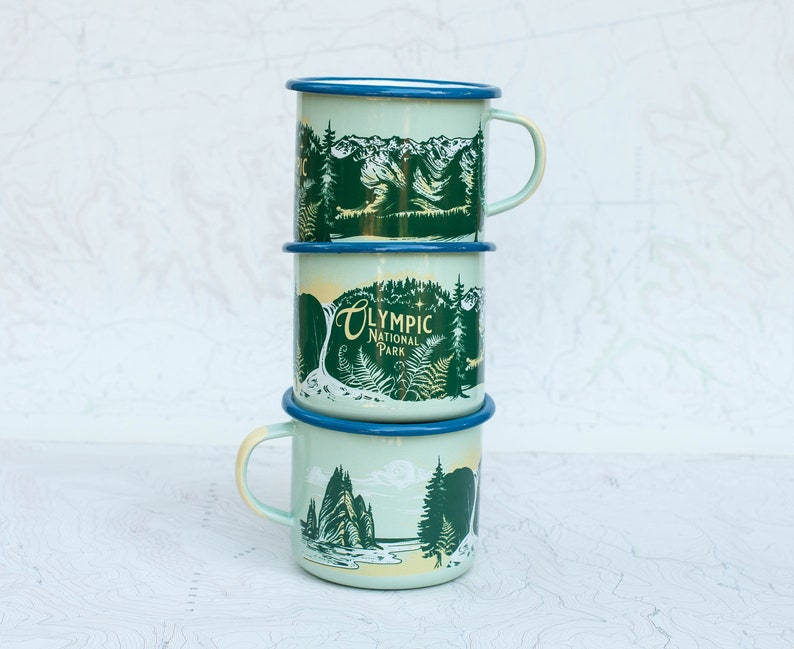 May include: A stack of three enamel camping mugs with a green and blue design. The top mug features a mountain scene, the middle mug says "Olympic National Park", and the bottom mug features a forest scene.