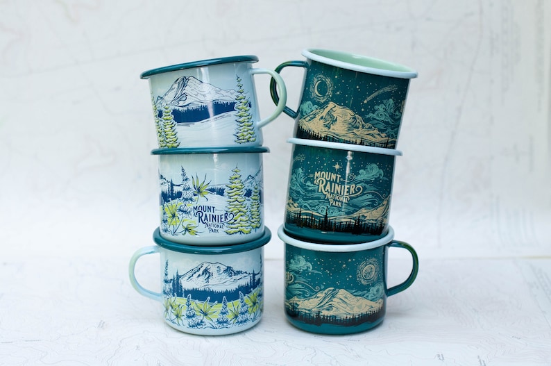May include: A stack of six enamel mugs with a blue and green color scheme. The mugs feature a mountain scene with the text "Mount Rainier National Park".