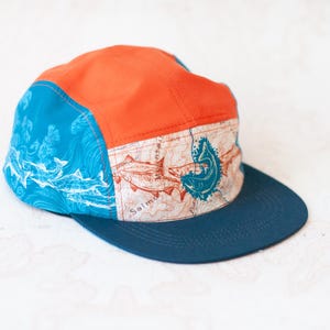 May include: A five-panel cap featuring a bright orange crown, a dark blue flat brim, and side panels with a blue and white fish pattern. The front panel displays a beige map print with salmon illustrations and the text Salmon River.