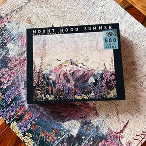 Pu&ograve; includere: Un puzzle da 500 pezzi con una vista estiva del Monte Hood. La scatola del puzzle mostra il testo "Mount Hood Summer" e un'opera d'arte della montagna, fiori selvatici e un cielo nuvoloso. Il puzzle parzialmente completato mostra una continuazione del design floreale.