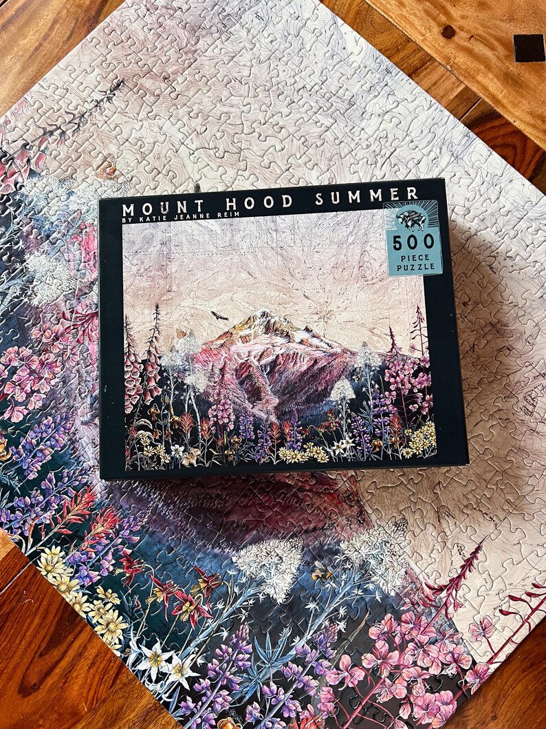 Pu&ograve; includere: Un puzzle da 500 pezzi con una vista estiva del Monte Hood. La scatola del puzzle mostra il testo "Mount Hood Summer" e un'opera d'arte della montagna, fiori selvatici e un cielo nuvoloso. Il puzzle parzialmente completato mostra una continuazione del design floreale.