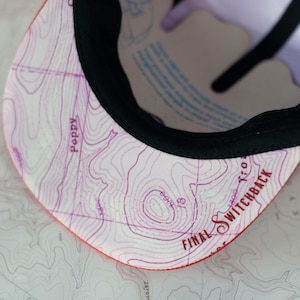 May include: A baseball cap with a map design on the underside of the brim. The map is in shades of purple and white, with the words "Poppy" and "Final Switchback" visible. The cap has a black brim.