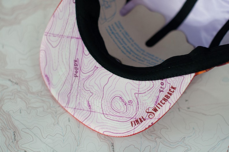 May include: A baseball cap with a map design on the underside of the brim. The map is in shades of purple and white, with the words "Poppy" and "Final Switchback" visible. The cap has a black brim.