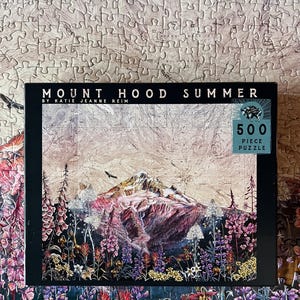 May include: A 500-piece jigsaw puzzle featuring a vibrant illustration of Mount Hood in summer. The puzzle box displays the title "Mount Hood Summer" and the artist's name. The image showcases a colorful landscape with flowers and a mountain.