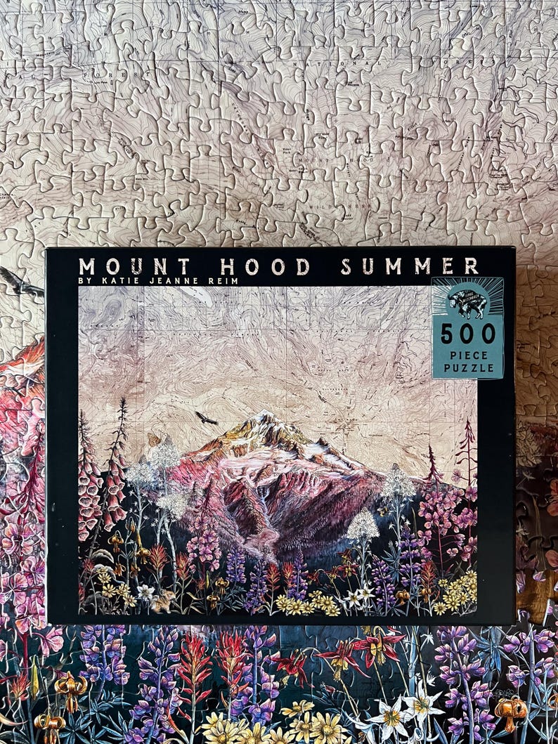Pu&ograve; includere: Un puzzle da 500 pezzi con una vivace illustrazione del Monte Hood in estate. La scatola del puzzle mostra il titolo "Mount Hood Summer" e il nome dell'artista. L'immagine mostra un paesaggio colorato con fiori e una montagna.