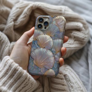 Puede incluir: Una funda de teléfono con un diseño de conchas marinas en tonos azules, rosas y dorados. La funda tiene un aspecto texturizado con un patrón repetitivo de conchas marinas. La funda del teléfono se sostiene en una mano.