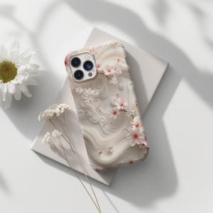 Könnte beinhalten: Eine weiße Handyhülle mit Blumenmuster. Die Hülle hat einen weißen Hintergrund mit rosa Blumen und einem weißen Wirbelmuster. Die Handyhülle hat einen Ausschnitt für das Kameraobjektiv.