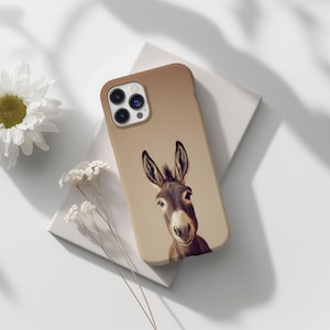 May include: A tan phone case featuring a detailed illustration of a donkey's face. The case has a smooth finish and a camera cutout. The donkey has brown and white fur, with large ears. The case is resting on a white surface with flowers.