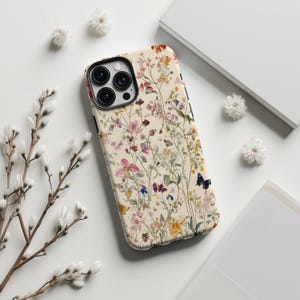 May include: A white iPhone case with a floral pattern featuring pink, red, yellow, and blue flowers and green leaves. The case has a black camera lens cutout and a black butterfly design.
