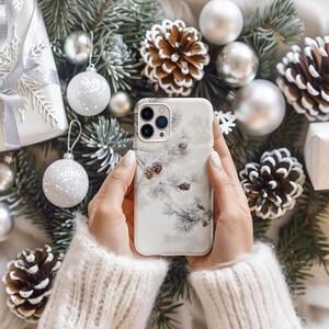 May include: A white phone case with a wintery design featuring pine branches and pine cones. The case is being held by a person wearing a white sweater.