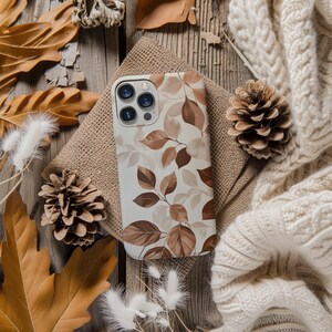 Op de afbeelding: Een witte telefoonhoes met een bruin en beige bladerpatroon ligt op een jute doek. De afbeelding toont herfstbladeren, dennenappels en een crèmekleurige gebreide deken, wat een herfstthema creëert.