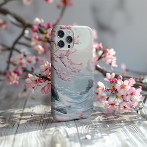 Peut inclure: Une coque de téléphone iPhone blanche avec un motif de fleurs de cerisier roses et un paysage bleu et blanc. La coque a une découpe pour l'objectif de l'appareil photo et un fond blanc.
