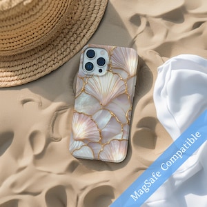 May include: A phone case with a seashell design. The case features a pattern of light pink and white seashell shapes outlined with gold lines. The phone case is on a sandy surface with a straw hat and white fabric in the background. The text "MagSafe Compatible" is visible.