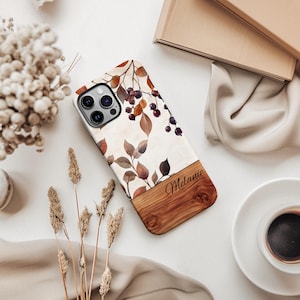 Puede incluir: Una funda de teléfono blanca con una base de aspecto de madera y un diseño floral con hojas y bayas marrones. El nombre "Melanie" está escrito en la parte de madera de la funda.