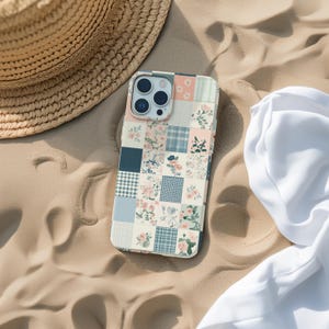 May include: A white phone case with a patchwork design featuring blue, pink and green squares with floral accents. The case has a camera lens cutout and a white background.