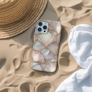Puede incluir: Una funda de teléfono con diseño de conchas marinas. La funda presenta un patrón de formas de conchas marinas blancas y rosas delineadas con líneas doradas. Está sobre arena junto a un sombrero de paja y tela blanca.