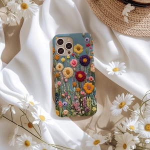 Pode incluir: Uma capa de telefone azul-petróleo adornada com um design floral bordado. A capa apresenta uma variedade de flores coloridas, incluindo flores amarelas, rosas e roxas, em um fundo de caules e folhas verdes. Margaridas e um chapéu de palha estão no fundo.