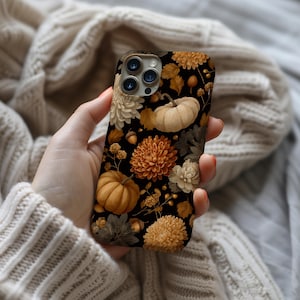 May include: A black phone case featuring a fall-themed design. The case is adorned with pumpkins, chrysanthemums, leaves, and acorns in shades of orange, cream, and brown. The floral and autumnal elements create a seasonal aesthetic.